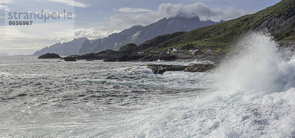 Wellen branden an die Felsenküste  dahinter Ferienhäuser und schroffe Berge  Flakstad  Lofoten  Nordland  Norwegen