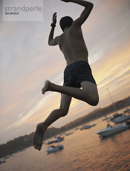 Ein Junge springt bei Sonnenuntergang an der Küste vom Dock ins Wasser.