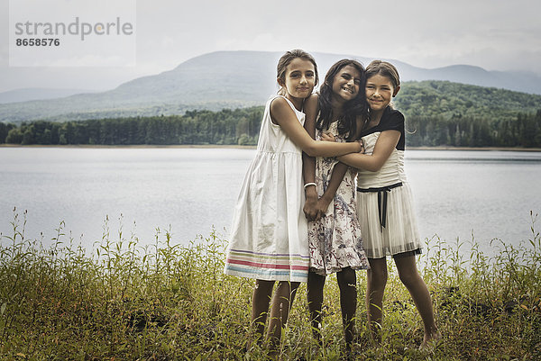 Drei junge Mädchen stehen am Ufer eines Sees und umarmen sich gegenseitig.