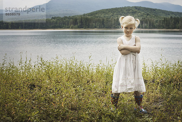 Ein junges Mädchen in einem weißen Sommerkleid  das mit verschränkten Armen steht und in die Kamera schaut.
