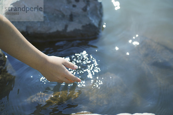 Ein junges Mädchen beugt sich vor  um ihre Hand in klares  flaches Seewasser zu halten.