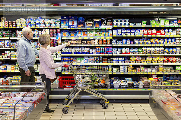 Seniorenpaar mit Einkaufswagen beim Einkaufen im Supermarkt  Kühlregal mit Molkereiprodukten  Deutschland