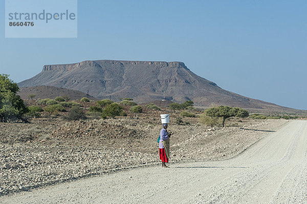 Frau mit einem Eimer auf ihrem Kopf steht an einer Schotterstraße in trockener Landschaft  Region Kunene  Namibia