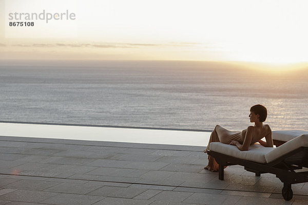 Frau im Kleid auf einem Sessel auf der Terrasse mit Blick auf das Meer bei Sonnenuntergang.