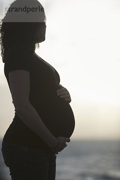 Silhouette einer hochschwangeren Frau  die am Strand steht und mit einer Hand auf ihrem Bauch ruht und mit der anderen den Bauch stützt.