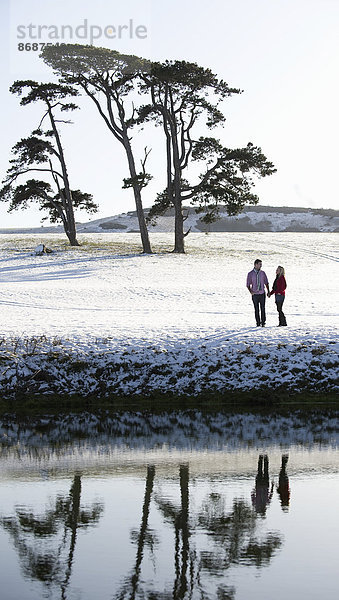 Ein Ehepaar steht auf einem schneebedeckten Feld an einem Fluss. Bäume im Hintergrund.