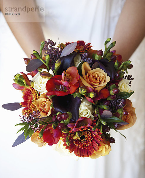 Eine Braut hält einen Brautstrauß aus bunten roten und orangefarbenen Blumen mit violett getönten Blättern.