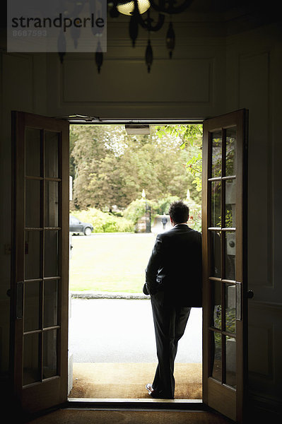 Silhouette eines Mannes  der in einer Türöffnung steht  sich gegen den Türrahmen lehnt und in den Garten blickt.