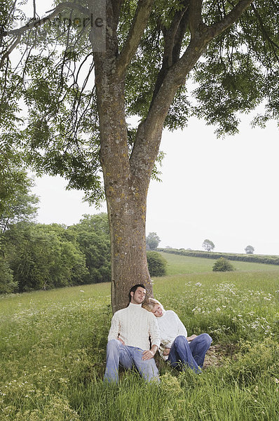 Ein Paar  das an einen Baum gelehnt auf einer Wiese sitzt.