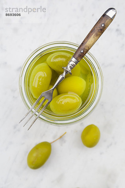 Einmachglas mit eingelegten grünen Oliven und einer Gabel auf weißem Marmor  erhöhte Ansicht