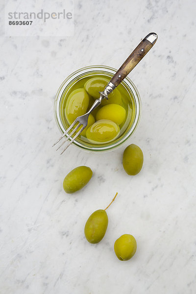 Einmachglas mit eingelegten grünen Oliven und einer Gabel auf weißem Marmor  erhöhte Ansicht