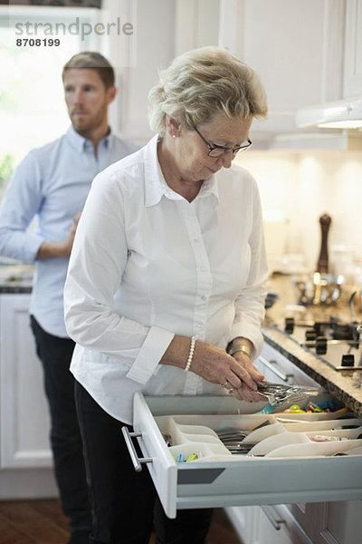 Seniorin arrangiert Besteck mit im Hintergrund stehendem Sohn in der Küche