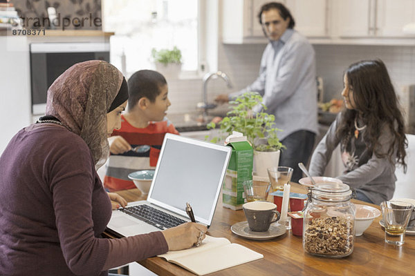 Muslimische Frau am Laptop mit Familie beim Frühstück in der Küche