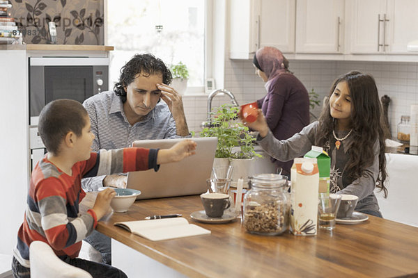 Muslimischer Mann arbeitet am Laptop in der Küche mit Familie beim Frühstücken