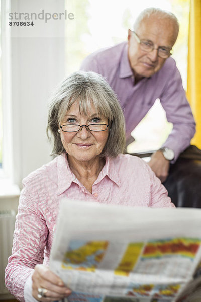 Porträt einer älteren Frau  die eine Zeitung mit einem Mann im Hintergrund zu Hause hält.