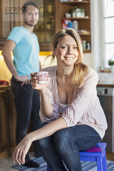 Porträt der Frau mit Kaffeetasse und Mann im Hintergrund in der Küche