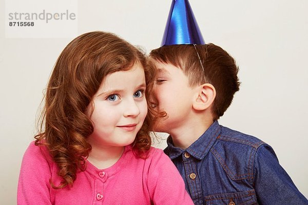 Junge trägt einen Partyhut und flüstert dem Mädchen zu.