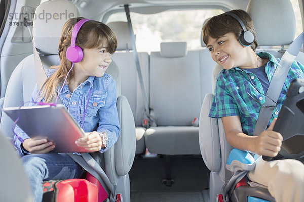 Glückliche Geschwister mit Kopfhörern und digitalen Tabletts auf dem Rücksitz des Autos