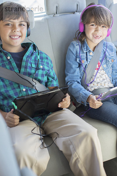 Portrait von glücklichen Geschwistern mit Kopfhörern und digitalen Tabletts auf dem Rücksitz des Autos