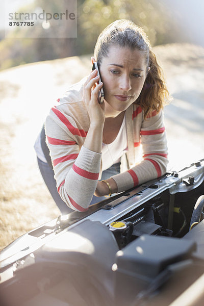 Frustrierte Frau spricht am Handy und schaut auf den Motor des Autos