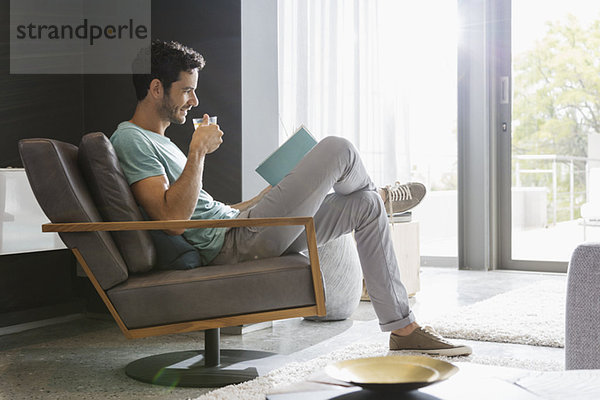 Mann trinkt Tee und liest Buch im Wohnzimmer