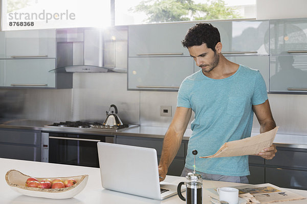 Mann mit Zeitung mit Laptop in der Küche