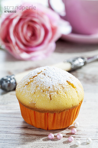 Muffin in Pappbecher  Rosenblüte  Tasse  Gabel und Zuckerperlen auf Holztisch