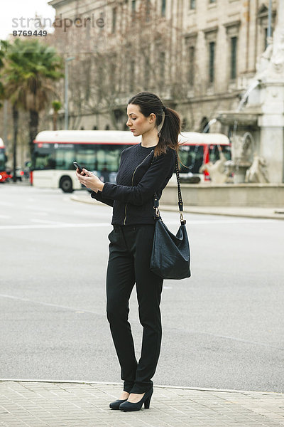Spanien  Katalonien  Barcelona  junge schwarz gekleidete Geschäftsfrau schaut auf ihr Smartphone vor einer Straße