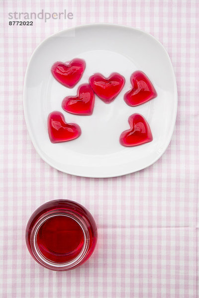 Teller mit roten Herzen in Form von Kirschgelee und Einmachglas mit Kirschgelee auf kariertem Tuch