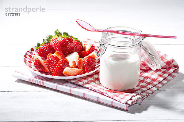 Glas Naturjoghurt mit Plastiklöffel  Teller mit geschnittenen und ganzen Erdbeeren (Fragaria) auf Küchentüchern und weißem Holztisch.