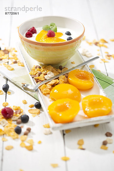 Schale aus laktosefreiem Joghurt mit Pfirsichstücken  Himbeeren  Heidelbeeren und Cerealien auf weißem Holztisch