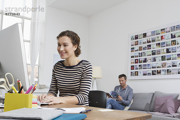 Frau zu Hause am Schreibtisch sitzend mit Computer und Mann im Hintergrund