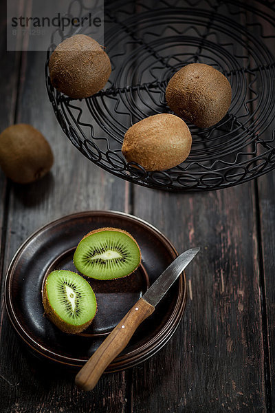 Drahtkorb und Teller mit ganzen und geschnittenen Kiwifrüchten auf Holztisch