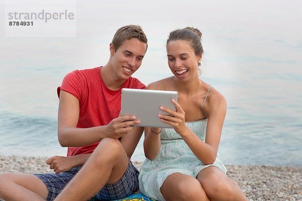 Bruder und Schwester sitzen am Strand und benutzen ein digitales Tablett.