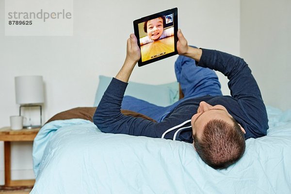 Mittlerer erwachsener Mann  der auf dem Bett liegt und ein digitales Tablett hochhält.