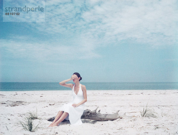 Frau auf Treibholz am Strand sitzend  die Augen mit der Hand bedeckend