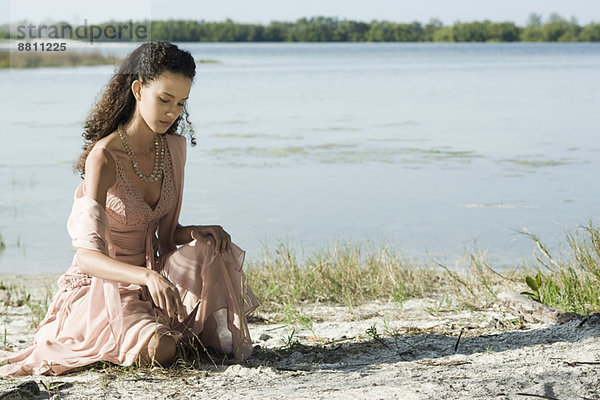 Junge Frau im Kleid am See sitzend  mit Stock in Sand gezeichnet