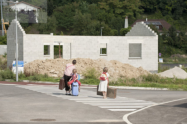 Mutter und Kinder gehen auf der Straße in Richtung unfertiges Haus  ziehen Koffer hinter sich her.