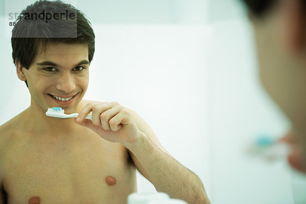 Junger Mann schaut in den Spiegel  hält die Zahnbürste  lächelt in die Kamera.
