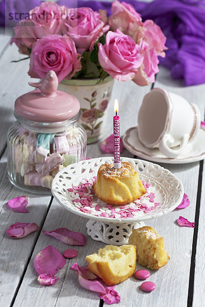 Muffin mit angezündeter Geburtstagskerze  Glas Marshmallows  Tasse und rosa Rosen auf Tisch