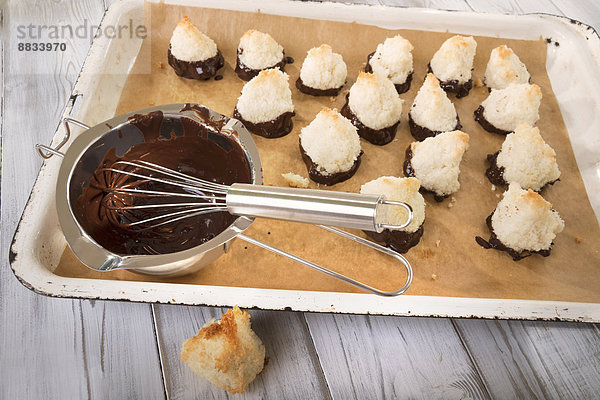 Backblech mit gebackenen Kokosmakronen und Topf mit flüssiger Schokoladenglasur