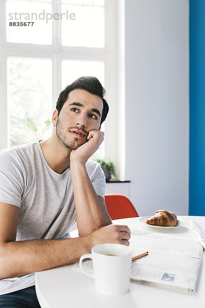 Porträt des nachdenklichen jungen Mannes am Frühstückstisch mit Zeitung