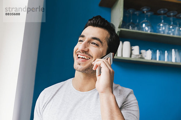 Porträt eines lachenden jungen Mannes  der mit einem Smartphone in seiner Küche telefoniert.