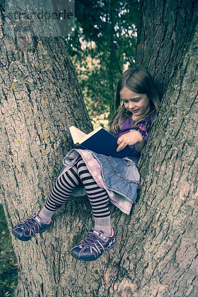 Kleines Mädchen sitzt auf einem Baumstamm und liest ein Buch.