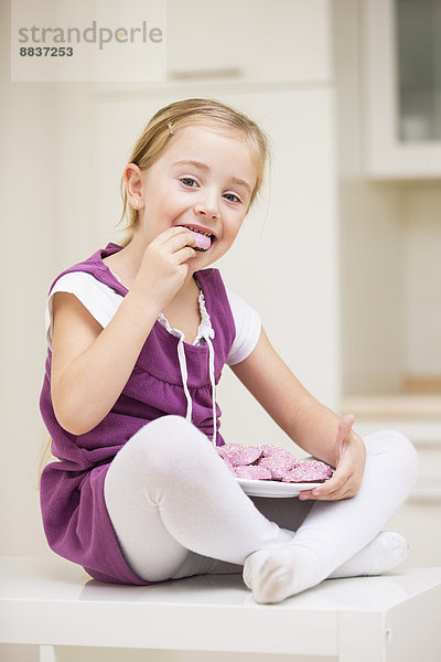 Porträt eines lächelnden kleinen Mädchens  das auf einem Tisch sitzt und rosa Kekse isst.