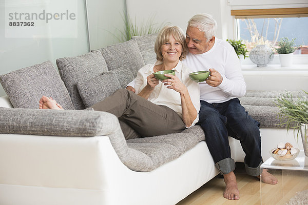 Seniorenpaar mit Teetassen nebeneinander auf Sofa im Wohnzimmer