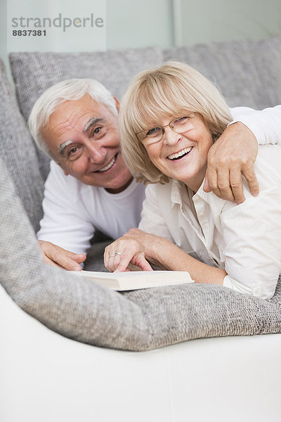 Porträt eines lächelnden Seniorenpaares mit nebeneinander liegendem Buch auf Sofa