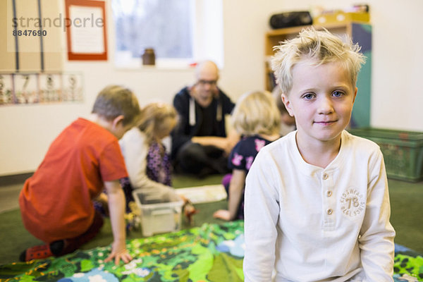 Portrait des Jungen mit Lehrerin und Klassenkameraden im Hintergrund im Kindergarten