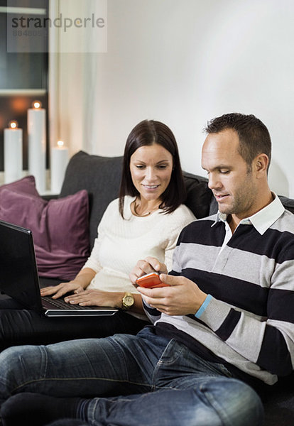 Paar mit Handy und Laptop auf dem Sofa zu Hause