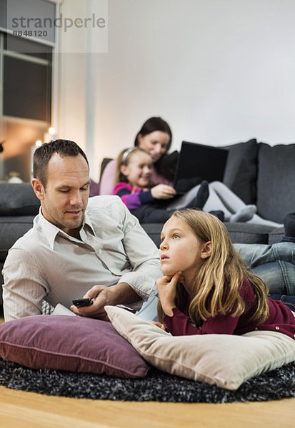 Vater und Tochter beim Fernsehen am Boden mit Familie im Hintergrund